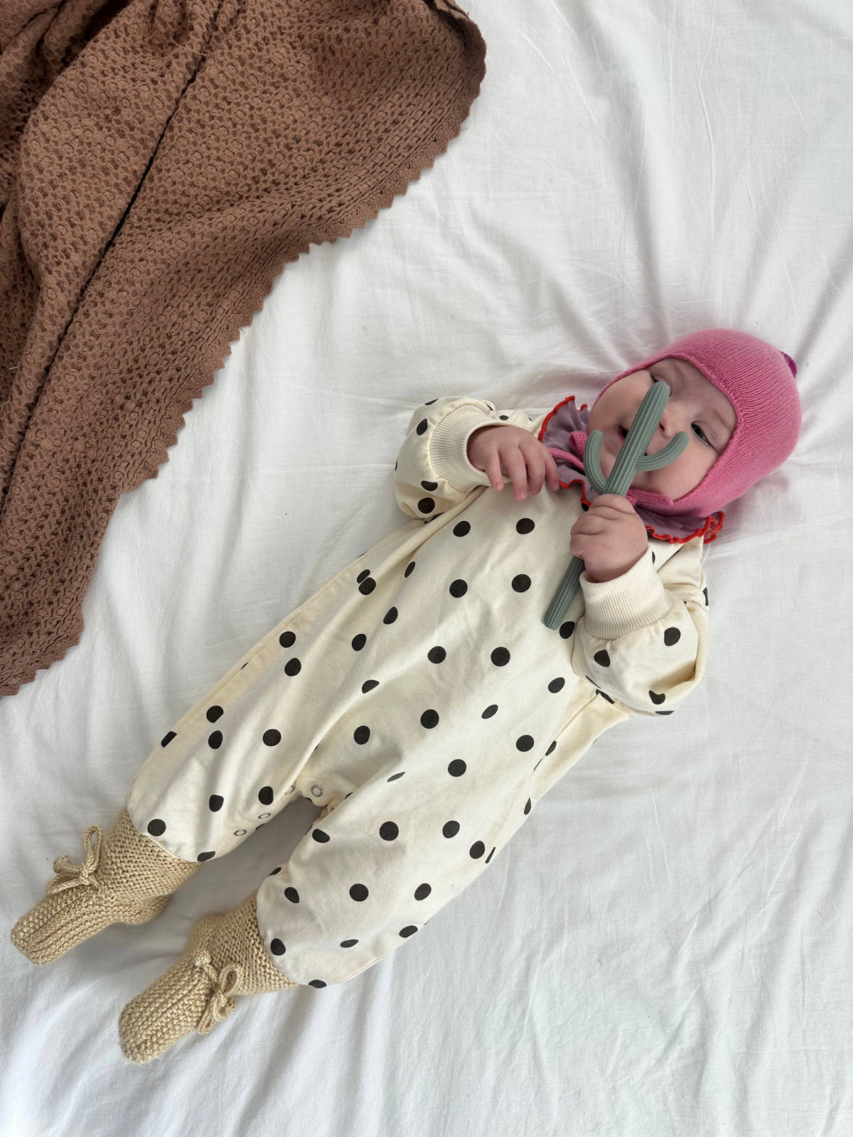 A baby lying on a white bed wearing a cream one-piece jumpsuit with brown polka dots, a pink knit hat, and beige knit booties, while holding a green toy.