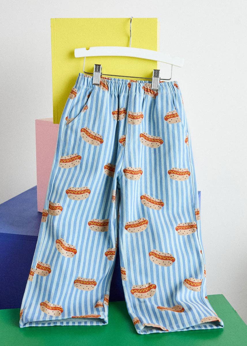 Light blue and white striped wide-leg pants with an all-over hot dog print, hanging on a white hanger against a colorful geometric background.