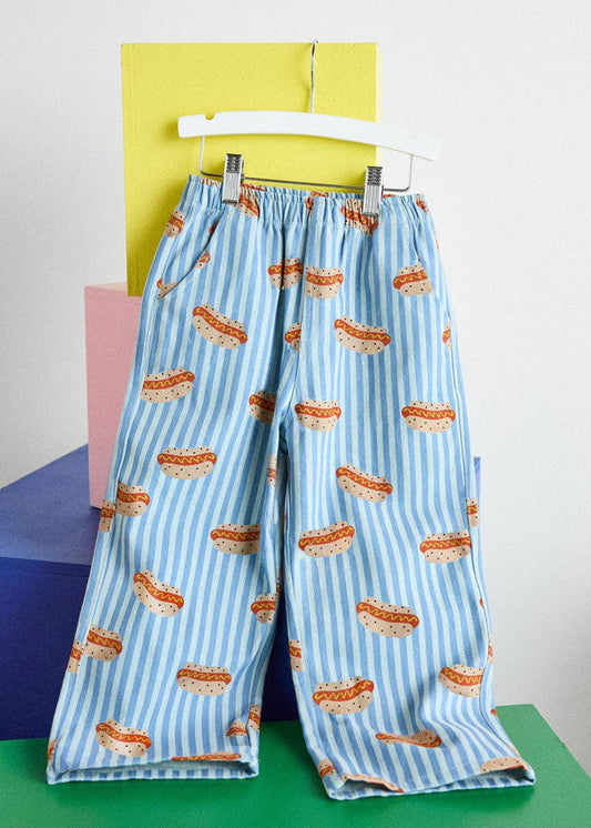 Light blue and white striped wide-leg pants with an all-over hot dog print, hanging on a white hanger against a colorful geometric background.