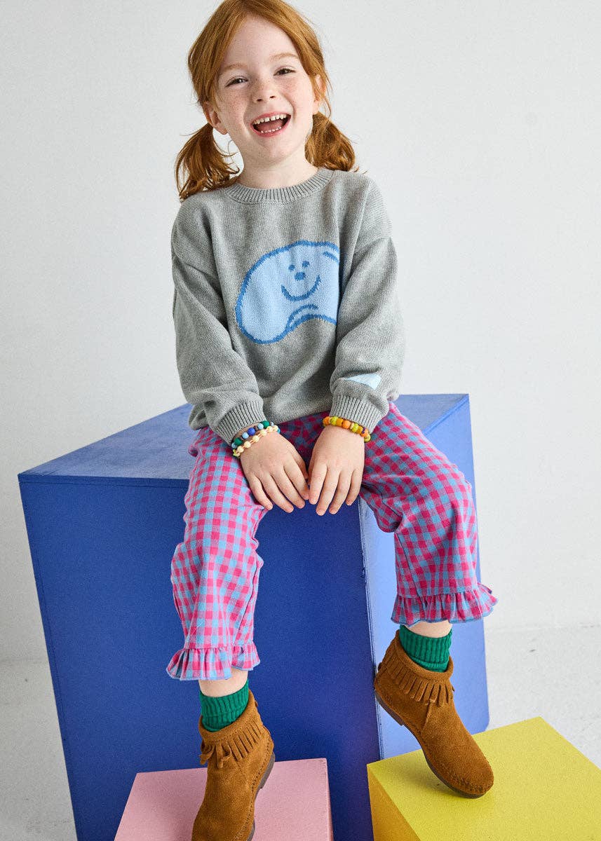 A young girl with red hair in pigtails sits smiling on colorful blocks, wearing a gray knit sweater with a blue smiley face, pink and white gingham pants, green socks, and brown suede boots.