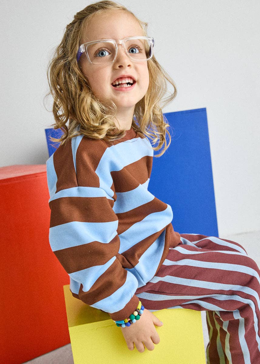 A young girl with blonde hair and clear glasses sits on a yellow block, wearing a brown and light blue striped long-sleeve top and matching striped pants. She is posed in front of colorful geometric blocks (red, yellow, blue) against a plain light gray wall.