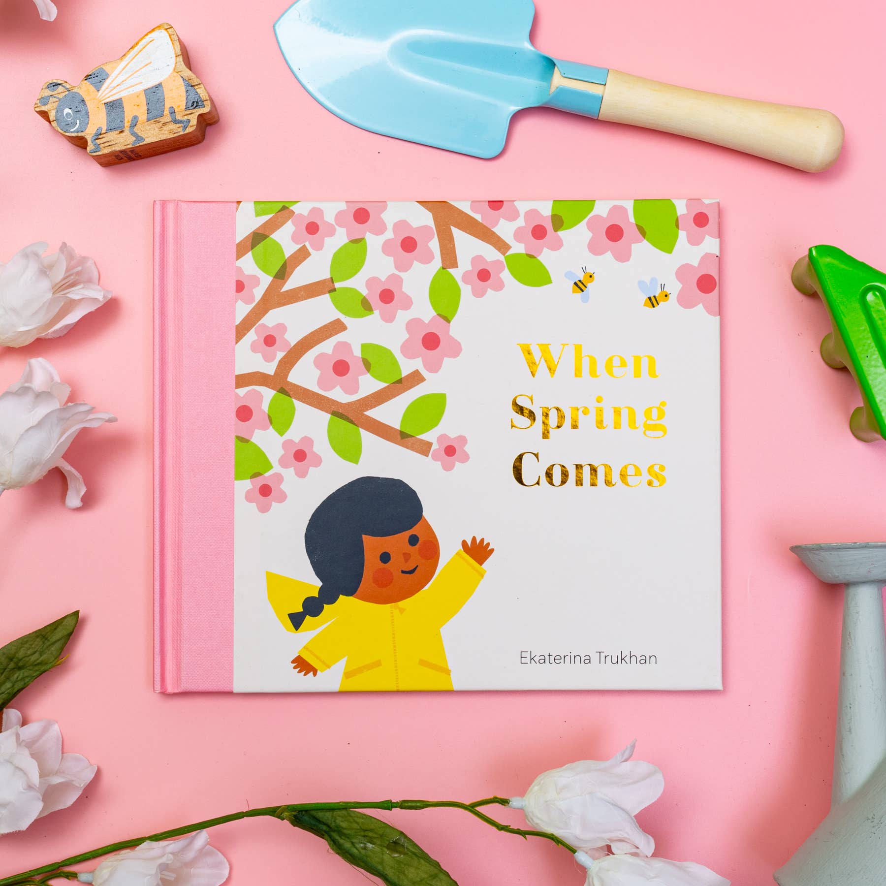 Children's book titled 'When Spring Comes' on a pink background with gardening tools and flowers.
