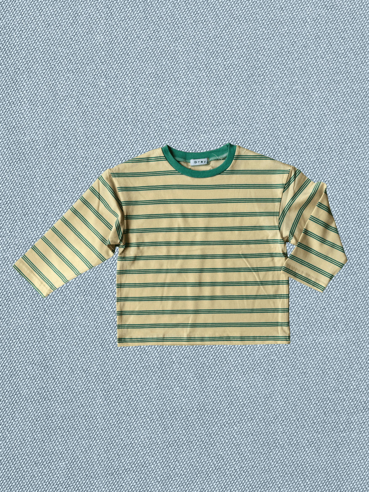 A long-sleeve striped shirt with a cream base and green horizontal stripes, featuring a solid green crew neck collar, laid flat on a textured blue background.