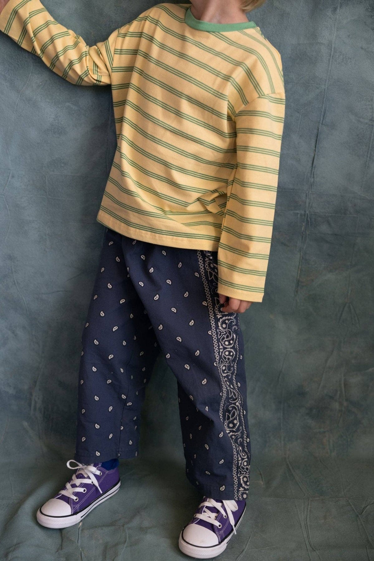 A child wearing a yellow and green striped long-sleeve shirt, navy blue pants with small white motifs and a paisley side stripe, and purple sneakers, standing against a textured blue backdrop.