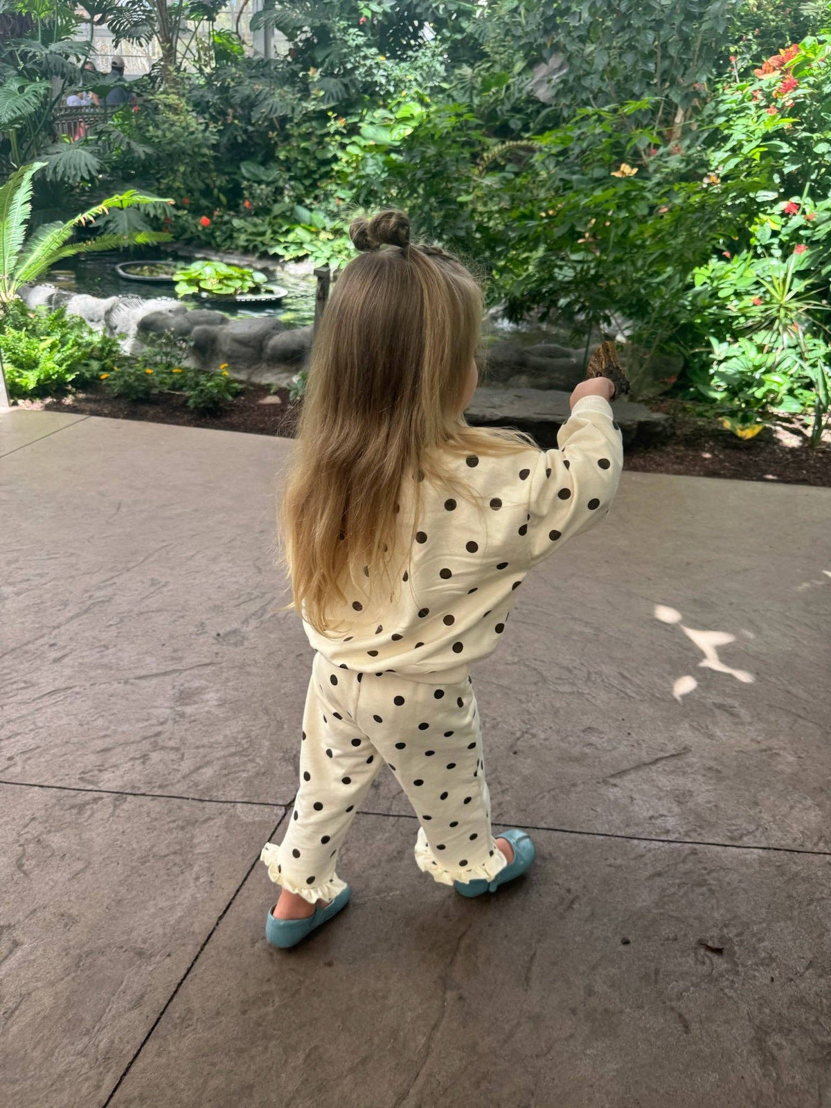 A young girl with long blonde hair in a high bun stands on a stone patio, viewed from behind, wearing a cream-colored pajama set with black polka dots and blue shoes, looking towards a lush garden background.
