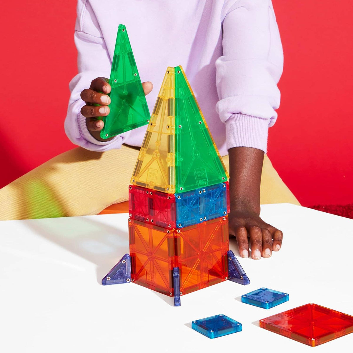 A boy in a lilac sweater is seated at a white tabletop and building a rocket ship with magnetic transparent blocks.
