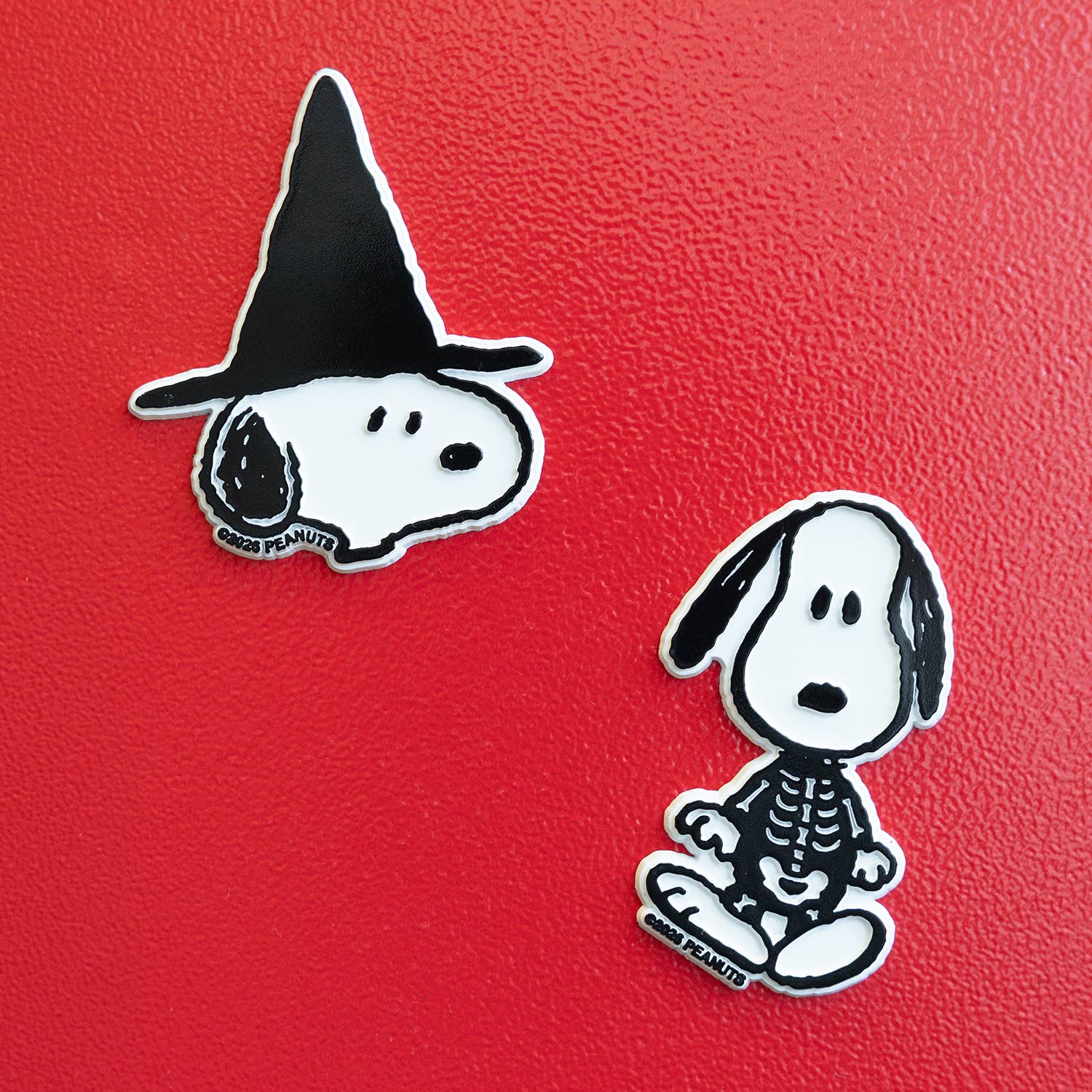 Two Snoopy Halloween-themed magnet on a textured red background. One pin shows Snoopy wearing a black witch hat, and the other shows Snoopy wearing a black skeleton costume.