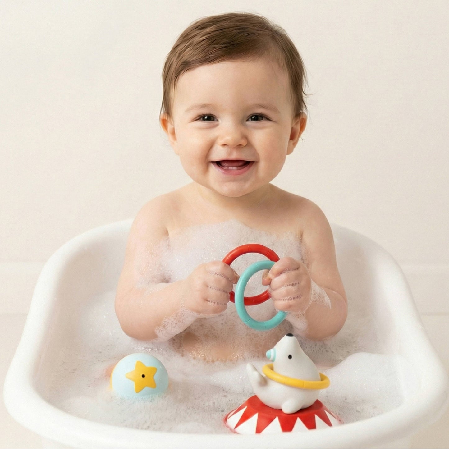 A smiling baby sitting in a white bathtub filled with soapy water, holding colorful toy rings. A white seal bath toy is visible in the tub.