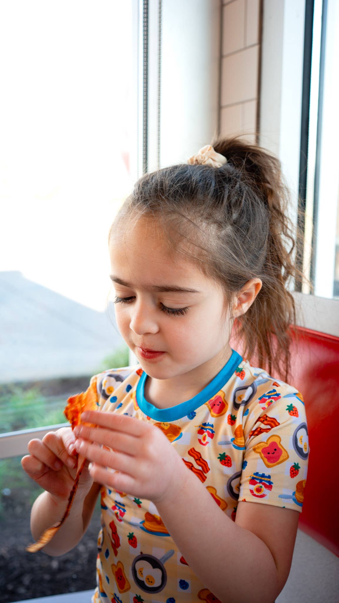 Young girl eating a slice of bacon indoors with a window in the background