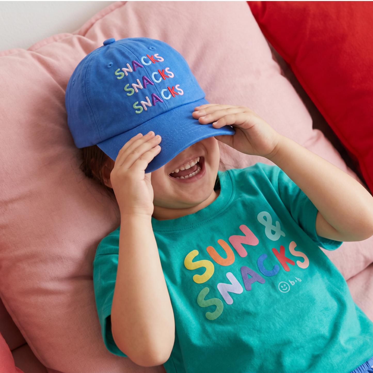 Child wearing a blue cap and teal shirt with colorful text, sitting on pink cushions.