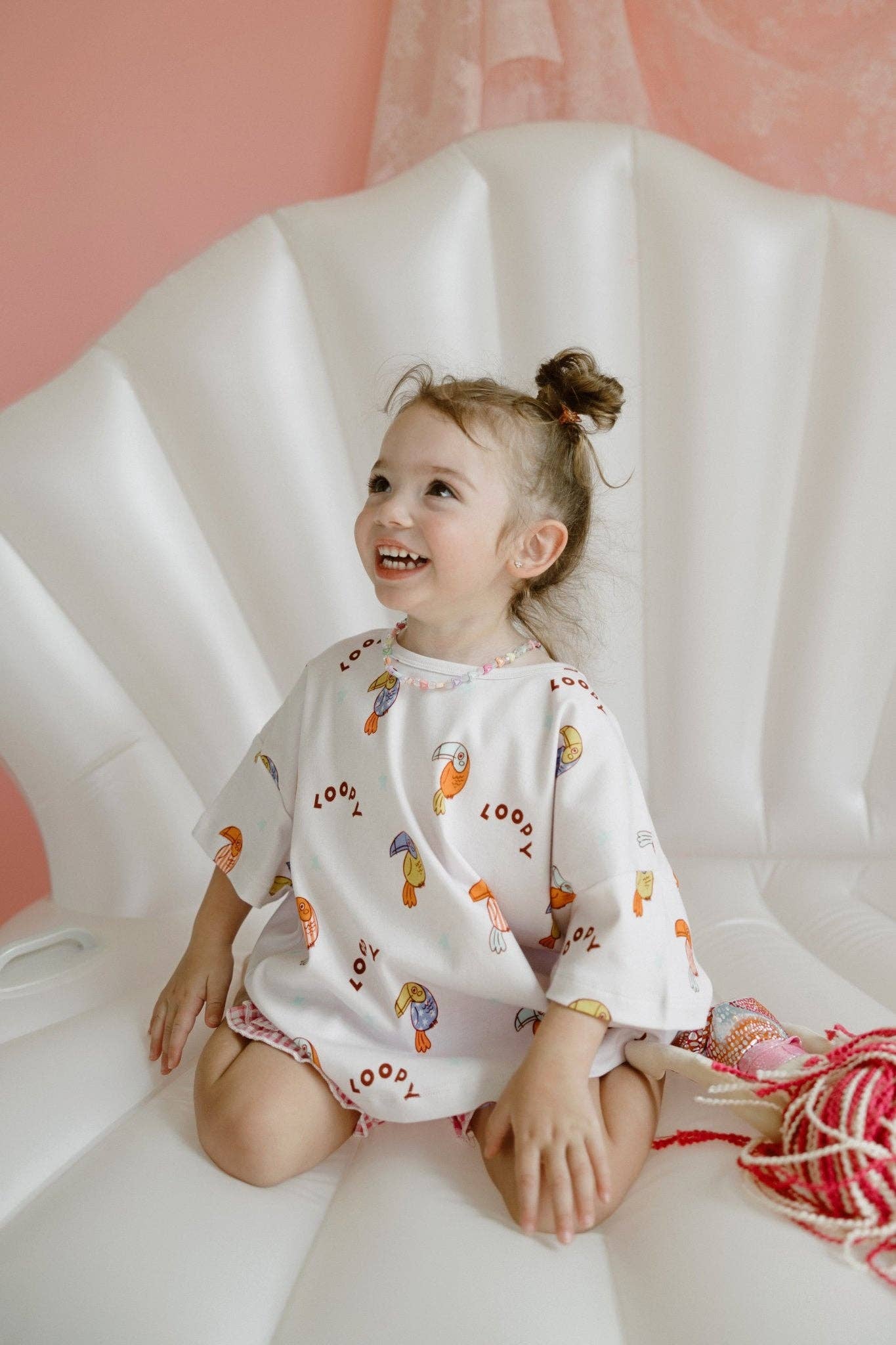 Child wearing a patterned outfit sitting on a white surface with a pink background