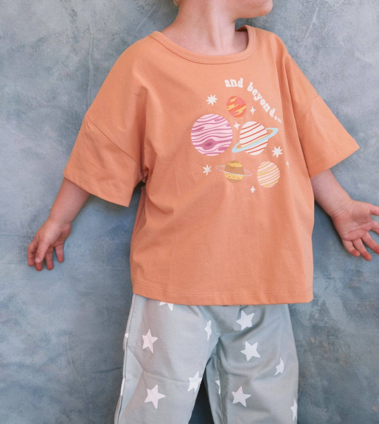 Child wearing an orange t-shirt with space-themed graphics and gray pants with star patterns against a gray background.