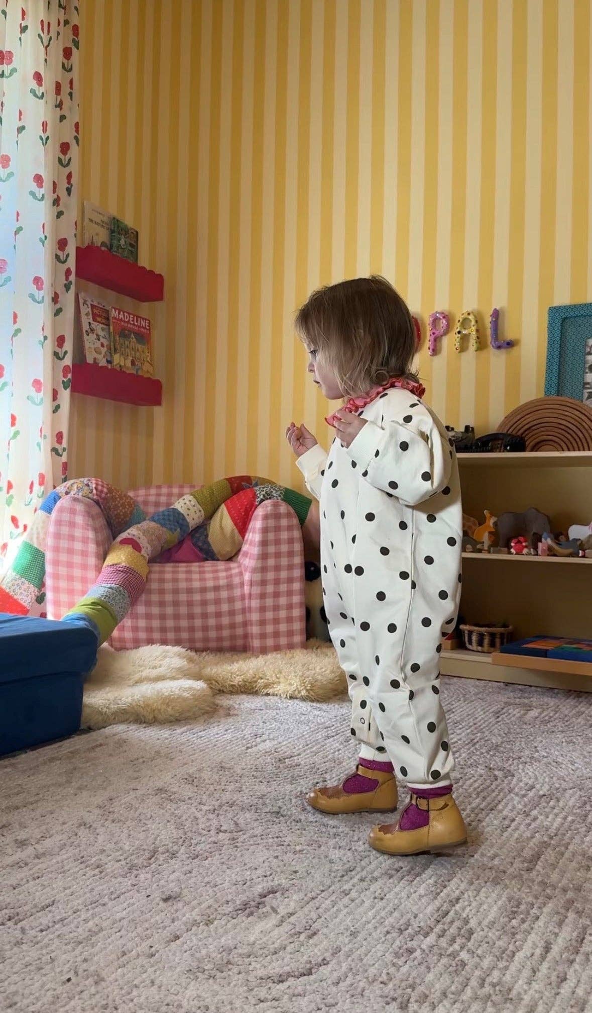 A toddler stands in a child's playroom wearing a white one-piece jumpsuit with brown polka dots, paired with purple socks and tan shoes. The room features yellow striped wallpaper, a pink-and-white checkered armchair, and a beige carpet.