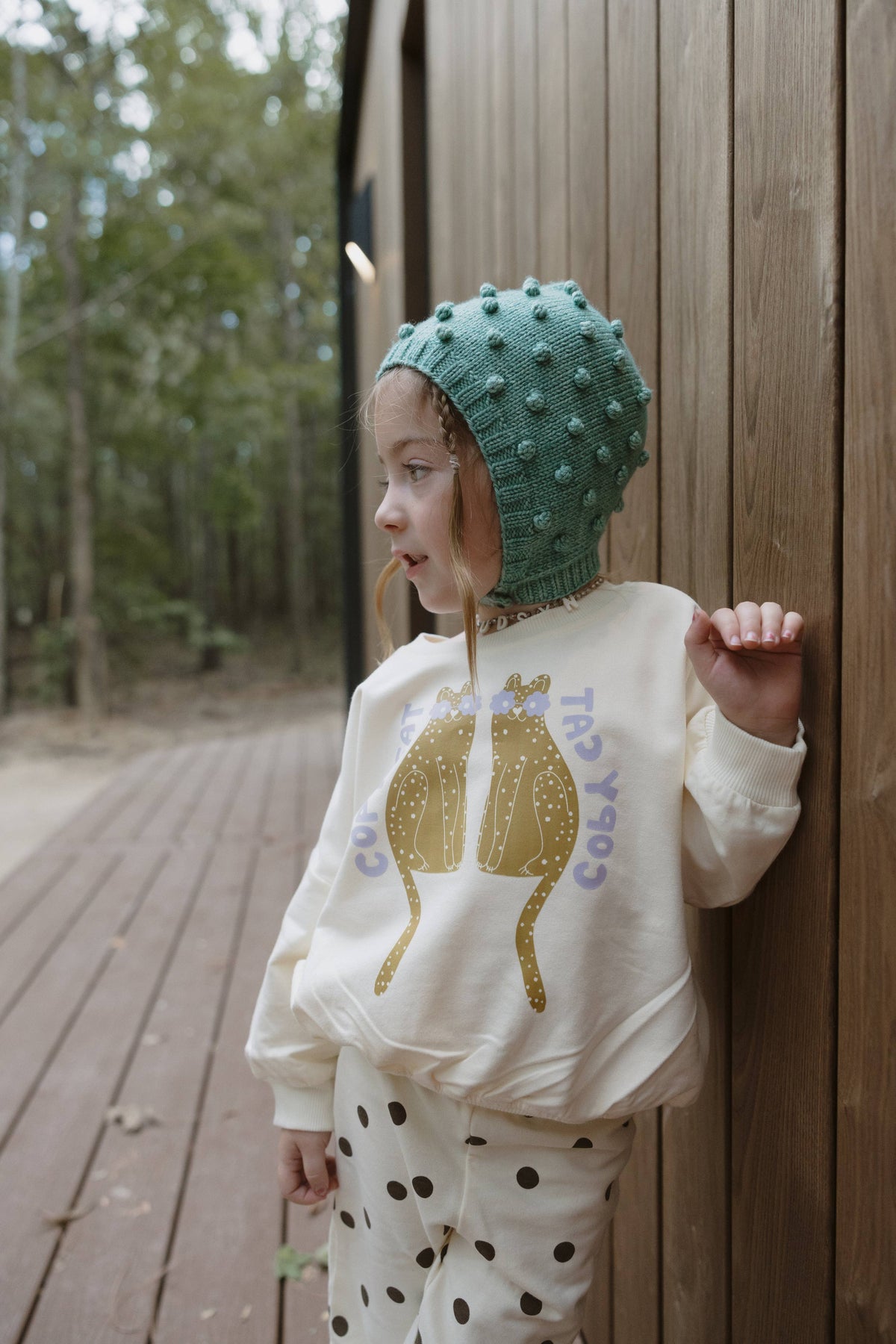 A young child wearing a cream-colored sweatshirt with a gold cat graphic and purple text, matching polka-dot pants, and a green textured bonnet, leaning against a wooden wall outdoors.