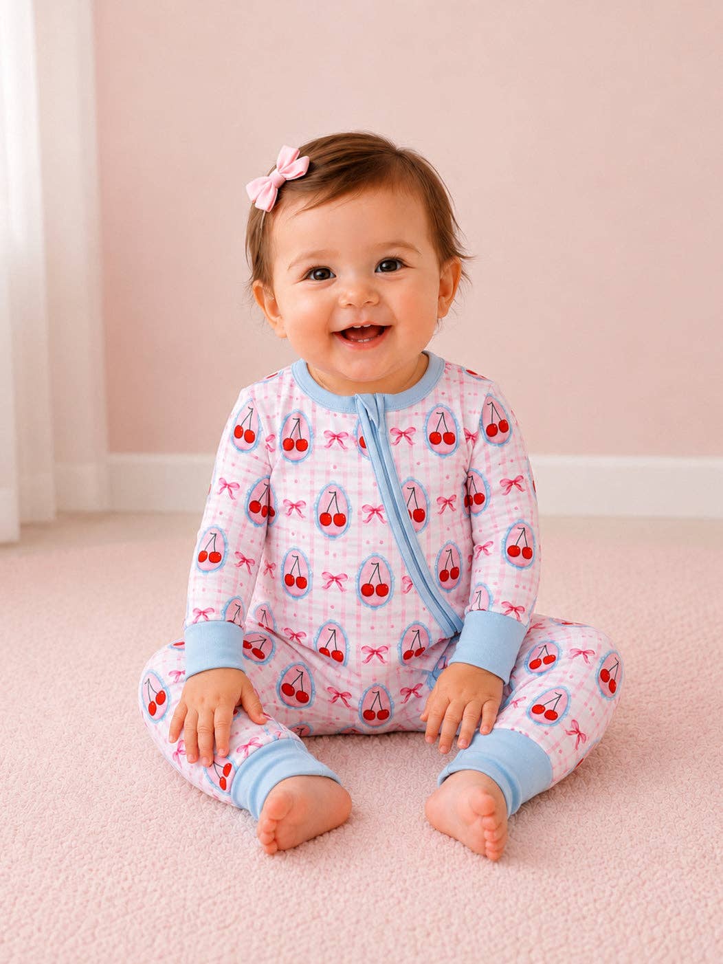 A baby girl sitting on a pink carpet wearing a light pink and white long-sleeve pajama set with a cherry print and light blue trim. She has a small pink bow in her hair.