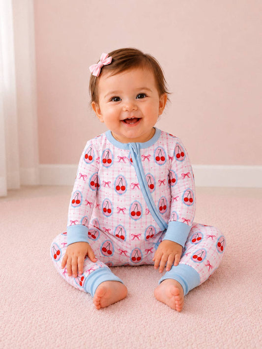 A baby girl sitting on a pink carpet wearing a light pink and white long-sleeve pajama set with a cherry print and light blue trim. She has a small pink bow in her hair.