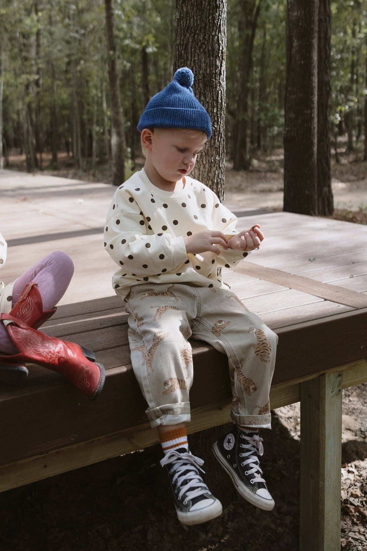 A toddler sits on a wooden deck in a wooded outdoor setting, wearing a cream long-sleeve top with brown polka dots, matching patterned pants, a blue knit pom-pom beanie, and black-and-white sneakers.