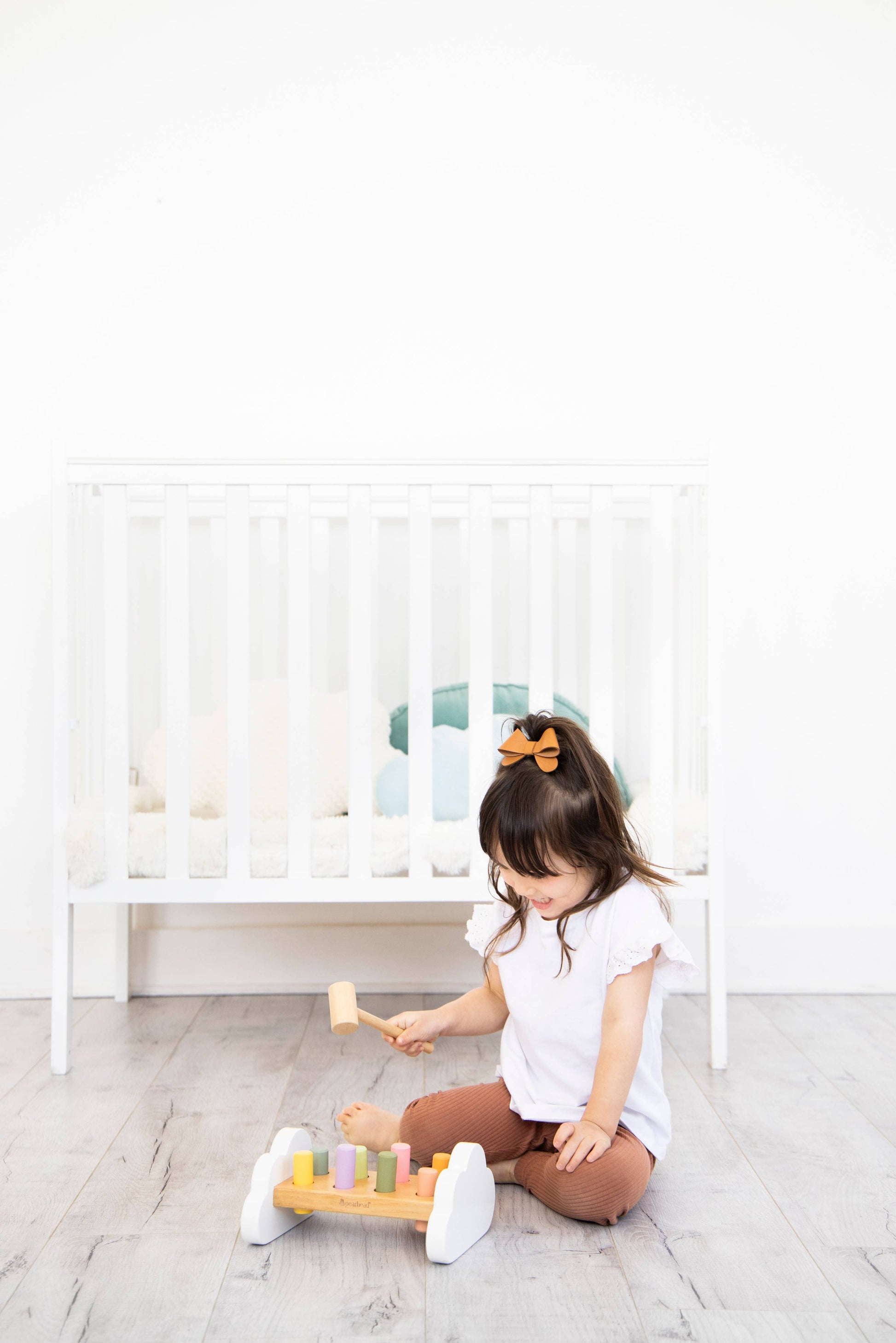 A young child sits on a wooden floor in front of a crib playing with a wooden hammer bench toy with colorful pegs and a wooden mallet.