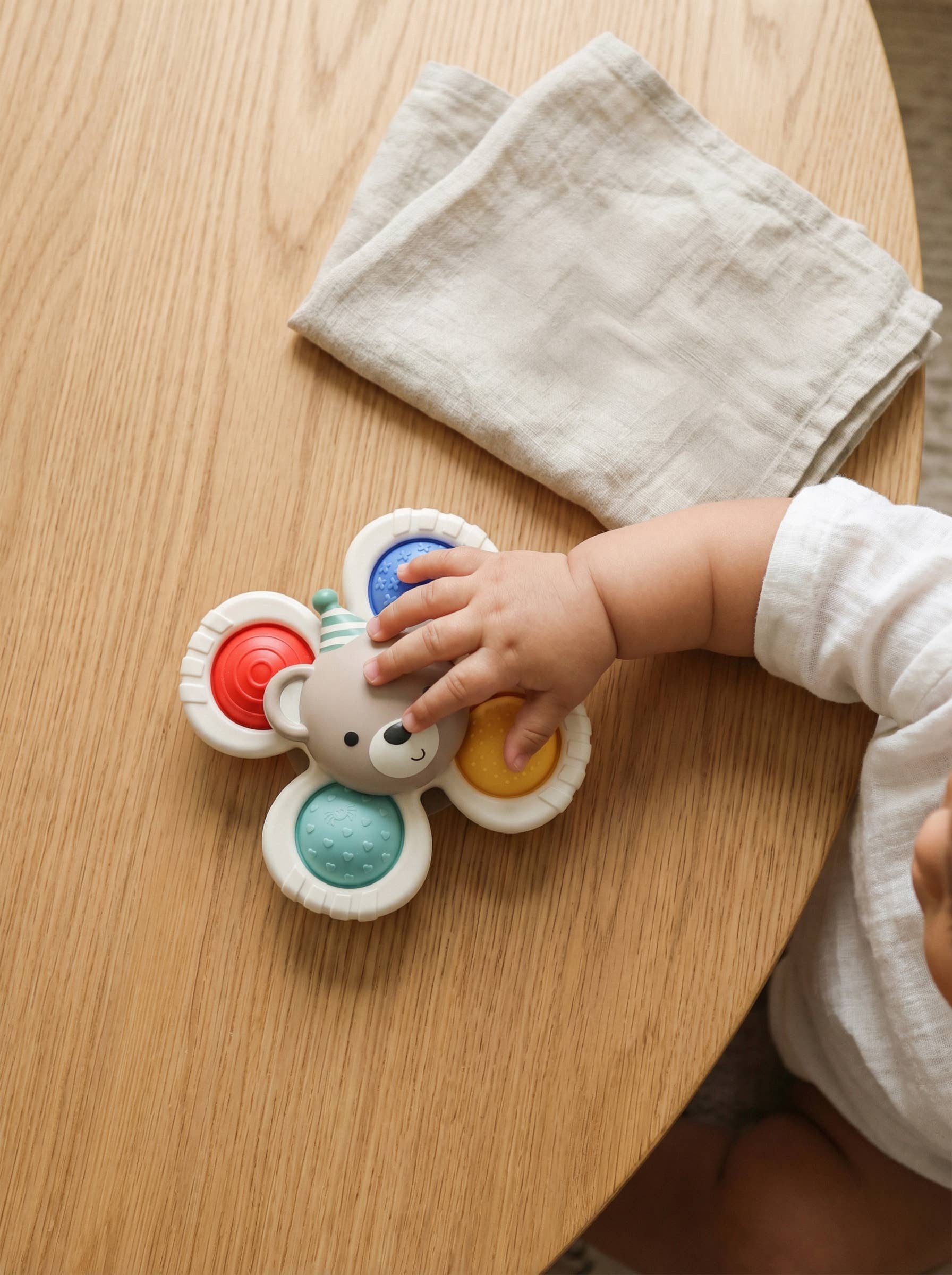 A baby's hand reaches for a flower-shaped sensory toy on a light wood table next to a folded beige cloth napkin.