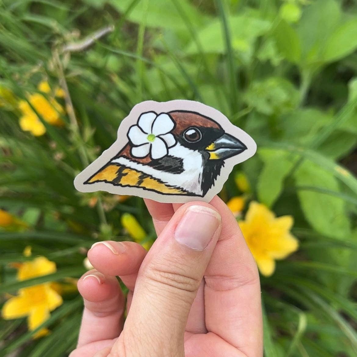 Hand holding a sticker of a bird with a flower on a green background with yellow flowers.