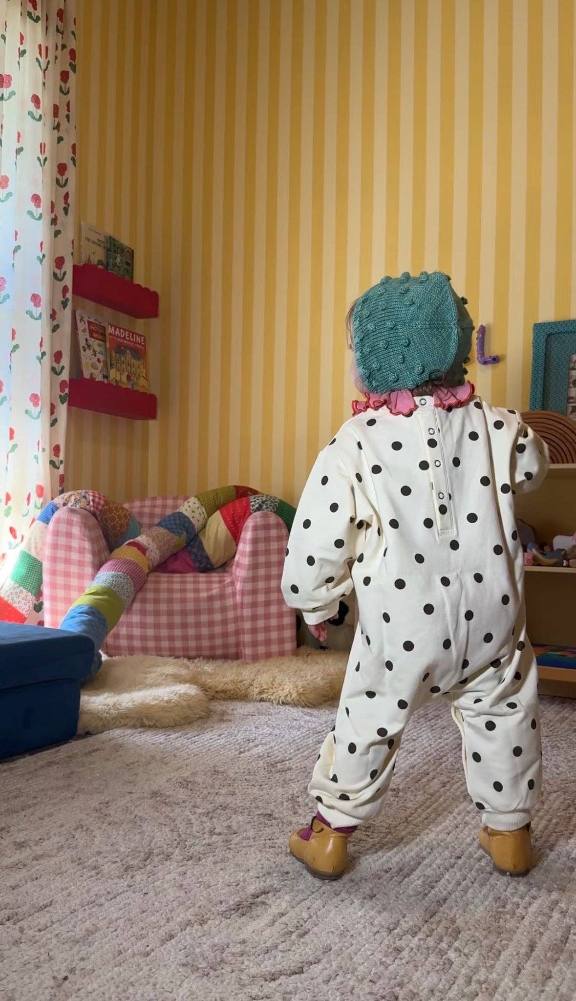 A baby wearing a cream-colored polka-dot jumpsuit, a teal knit hat, and tan booties stands in a child's playroom with yellow striped wallpaper.
