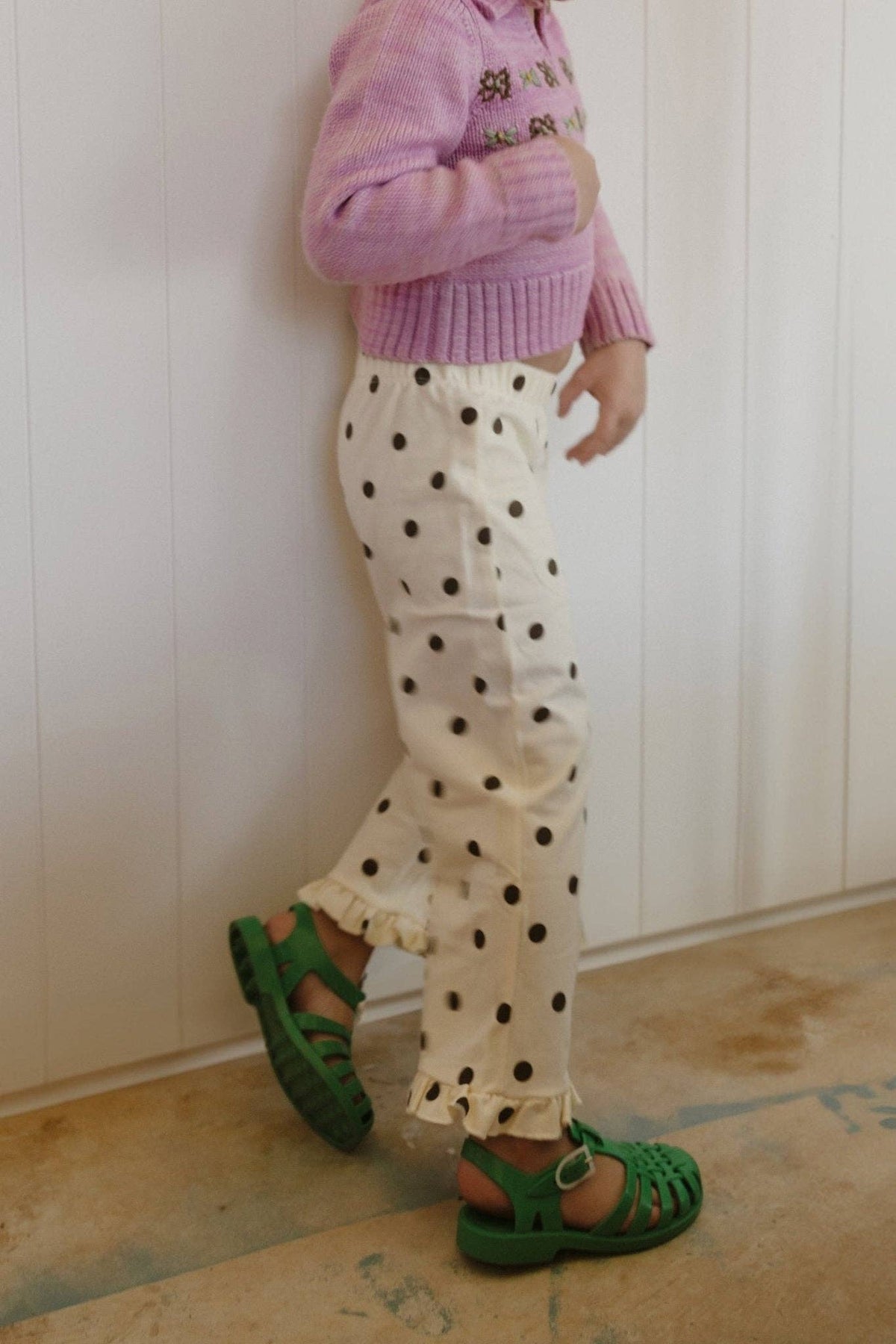 A child wearing cream pants with black polka dots, a pink knit sweater, and green sandals, standing against a white paneled wall.