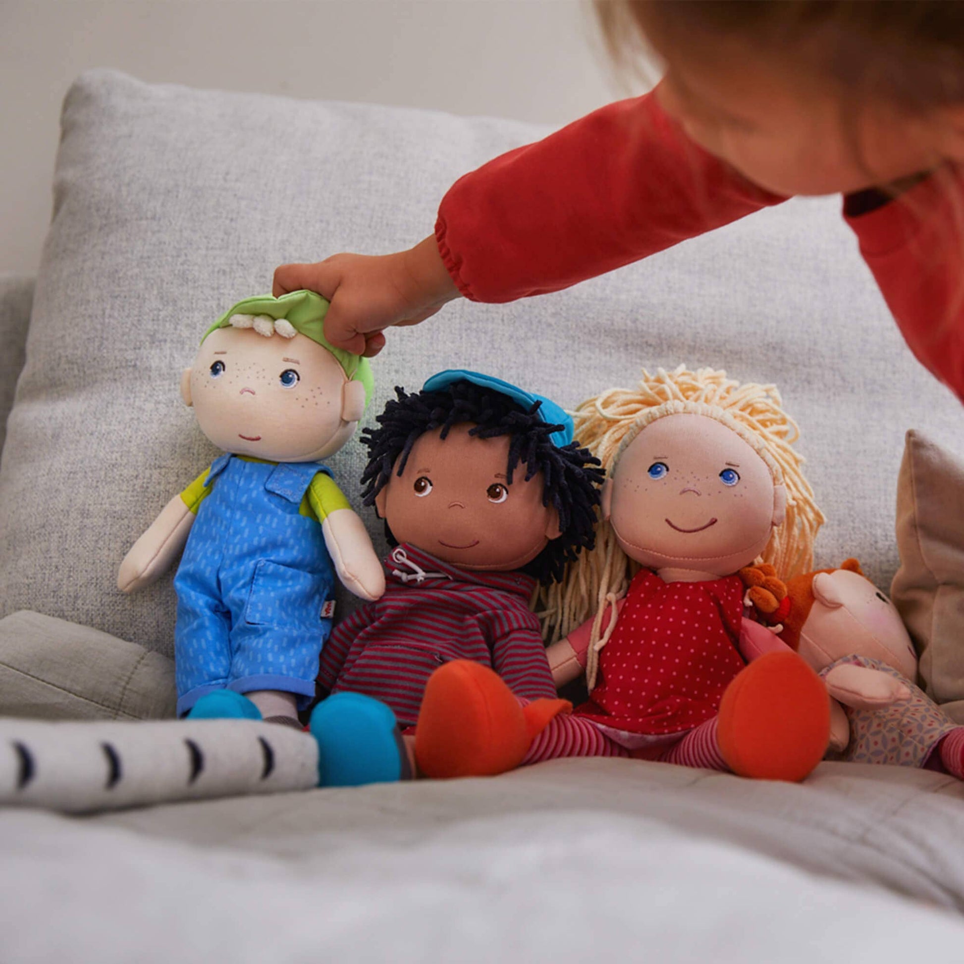 Children's dolls with different hair colors and outfits on a couch, with a child's hand reaching out.
