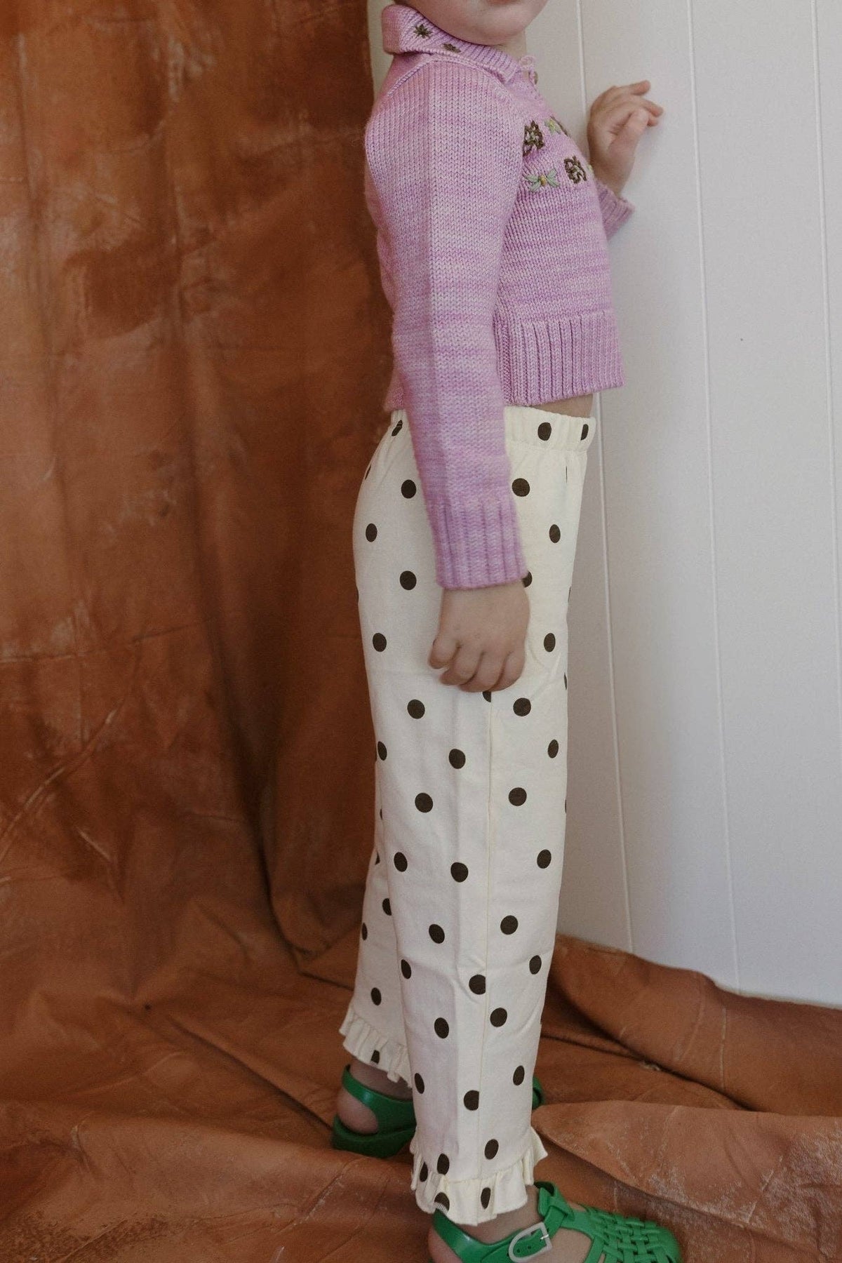 A child wearing a light purple knit cropped sweater and cream wide-leg pants with dark brown polka dots, standing against a split brown fabric and white paneled wall background.