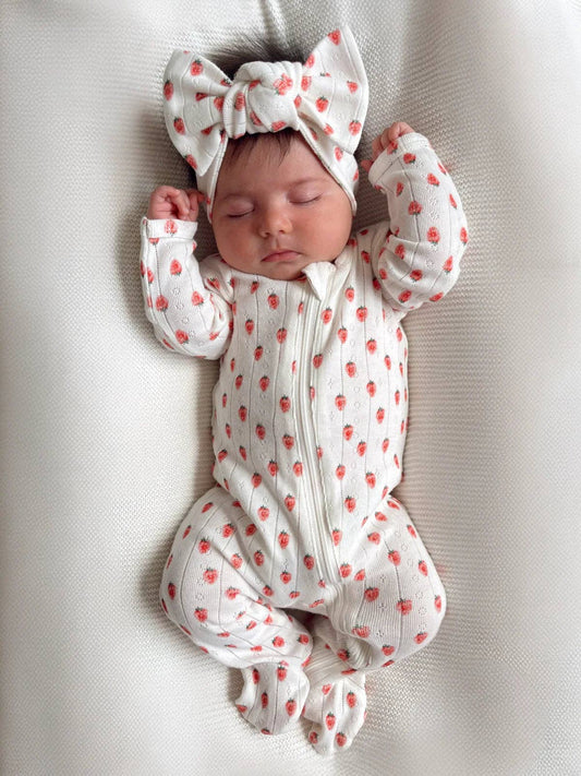 Newborn baby wearing a white outfit with red patterns and a matching headband, lying on a soft surface.