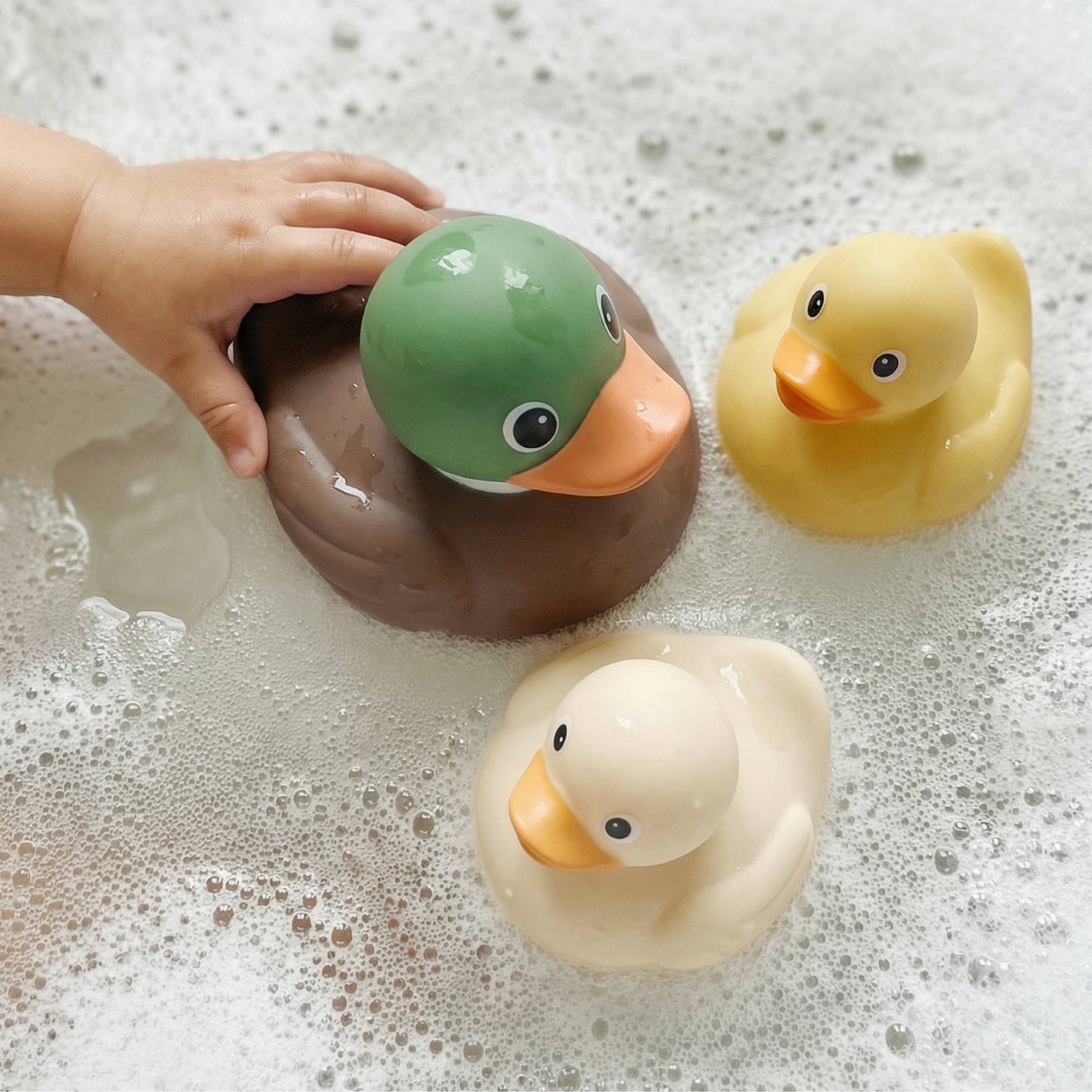 Three colorful rubber duck toys floating in bubbly soapy water, with a child's hand resting on the green duck.