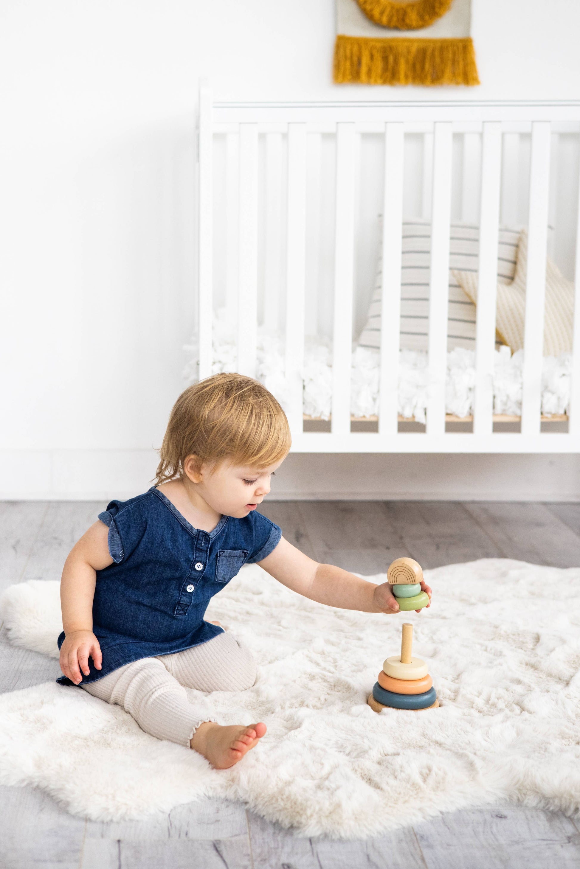 A child wearing a blue shirt is sitting on a fluffy carpet playing with a wooden stacking toy consisting of a peg with five multicolor earth tone rings and a wooden rainbow topper on a white background.