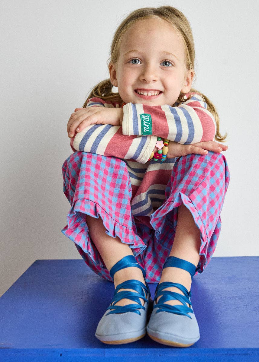 A young girl with blonde pigtails sits smiling on a blue platform against a white wall, wearing a pink and blue gingham dress, a striped long-sleeve top, and blue ribbon shoes.
