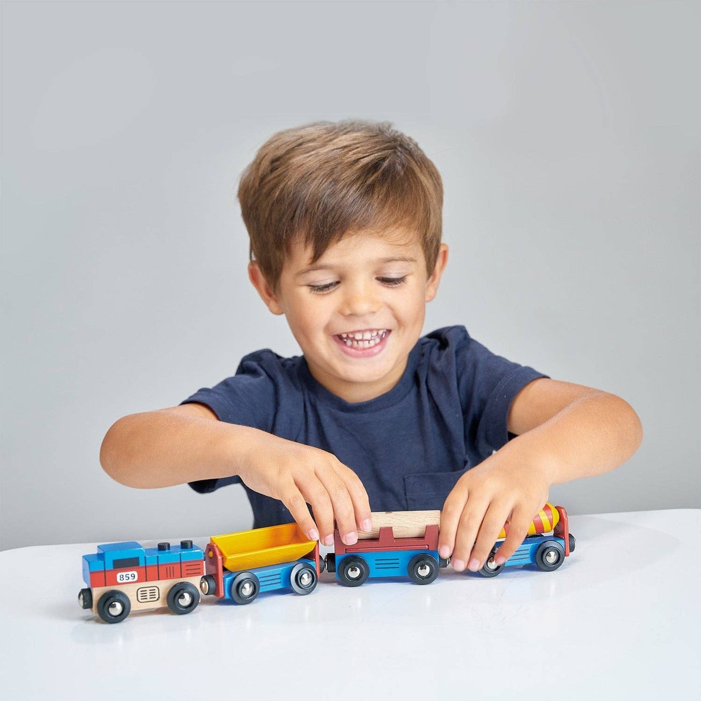 A child with a blue shirt is playing with a wooden train set with one engine and three colorful attached carriages designed to resemble industrial cargo goods.