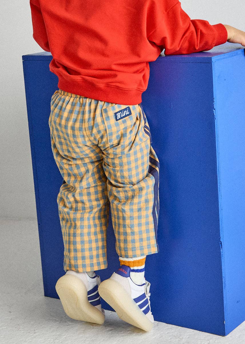 A child wearing yellow and beige checkered pants with side stripes, a red sweatshirt, and white sneakers, standing on a blue block.