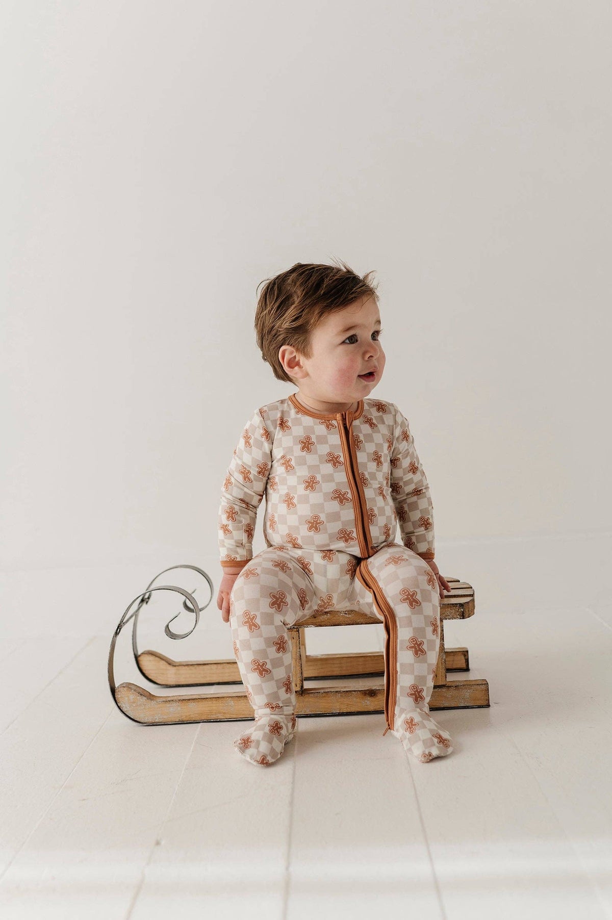A baby wearing gingerbread footie pajamas sitting on a sleigh in front of a white background.