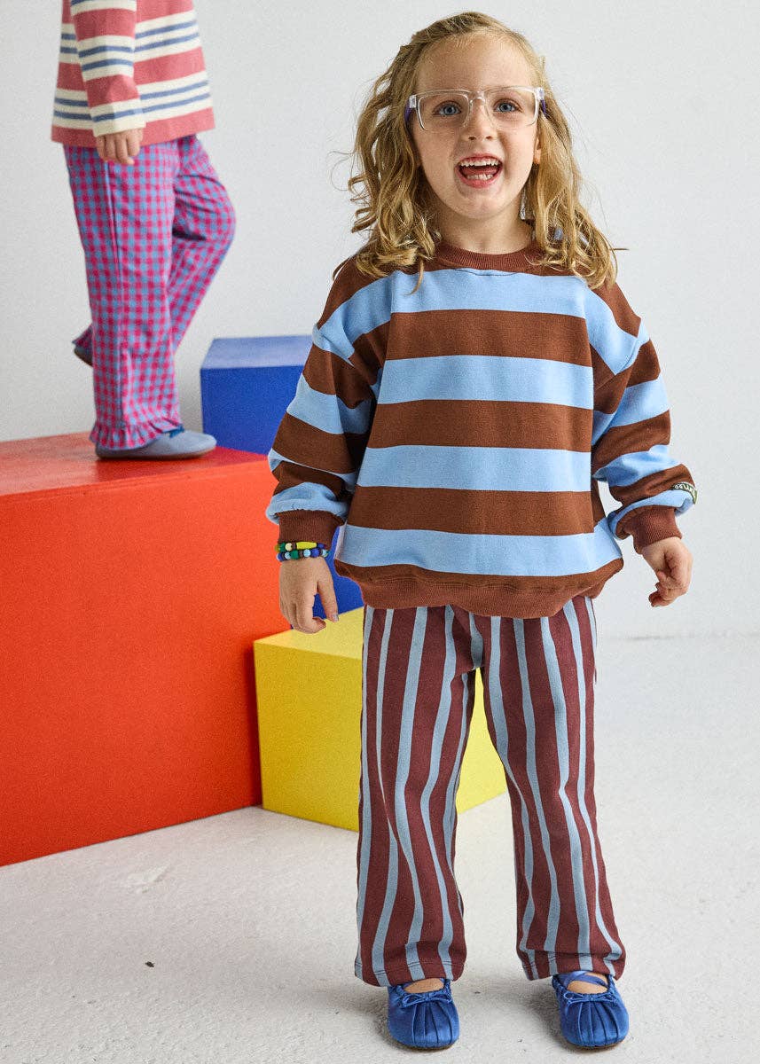 A young girl wearing a matching two-piece terry sweat set with brown and light blue horizontal stripes on the long-sleeve top and red and white vertical stripes on the pants, standing in front of colorful blocks.