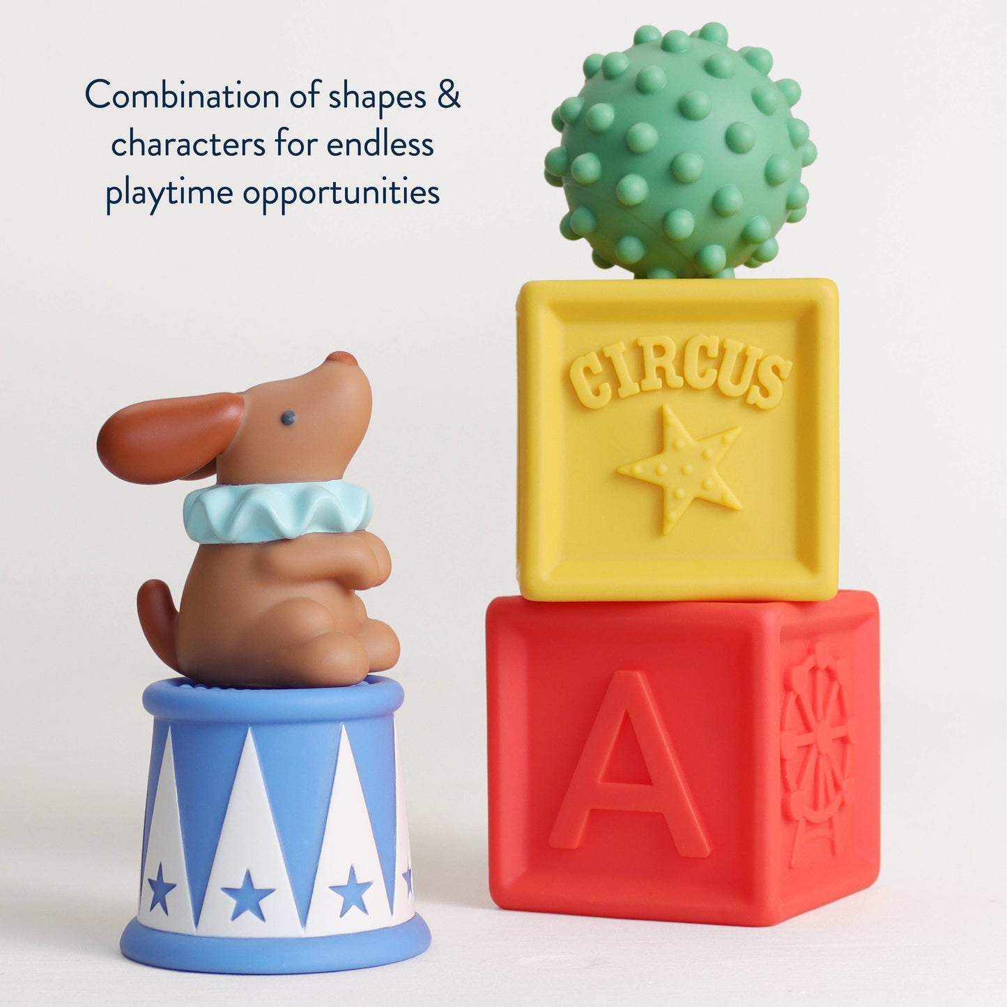 A children's toy set featuring a brown dog figurine wearing a light blue collar, sitting on a blue and white circus-themed base. Next to it is a stack of three embossed alphabet blocks in yellow, red, and blue, with a green bumpy ball toy on top.