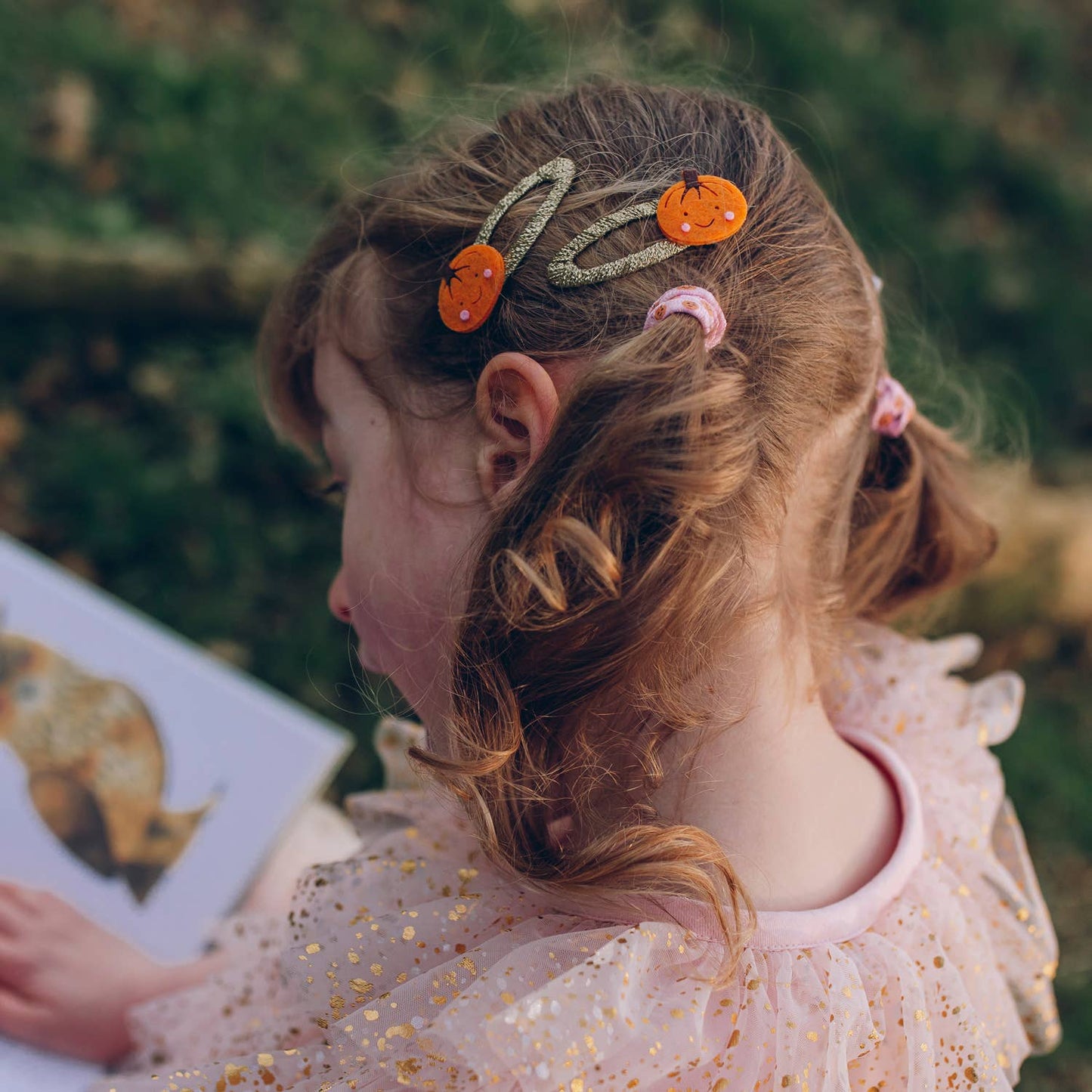 A young girl with styled brown hair wearing three pumpkin-shaped hair clips, photographed outdoors with a blurred green background.