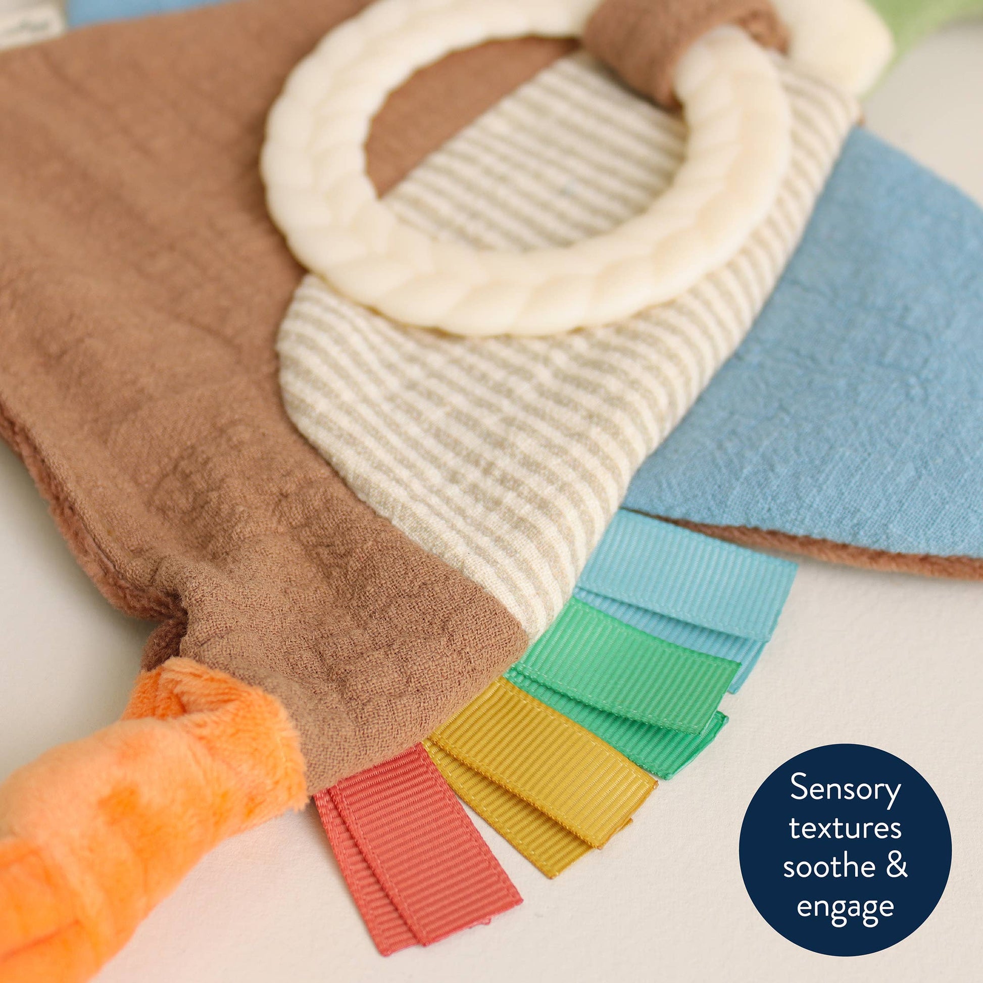 A close-up of a soft sensory toy with multiple textures and colors, including a brown fabric body, cream striped section, blue panel, orange plush pull, rainbow-colored ribbed tabs, and a cream ring handle. A dark blue circular text badge reads 'Sensory textures soothe & engage'.