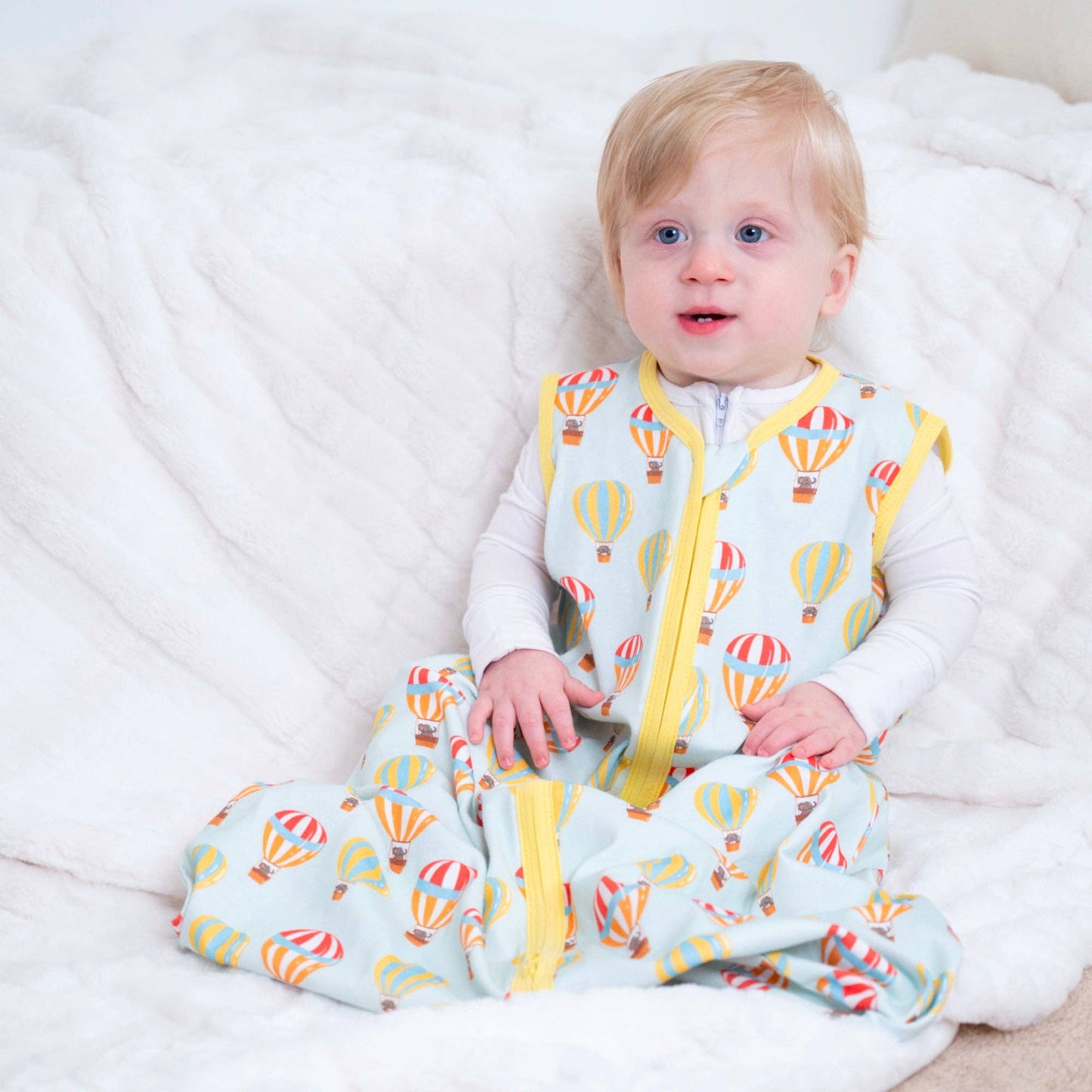 Baby wearing a colorful sleeping sack with hot air balloon pattern on a white blanket.