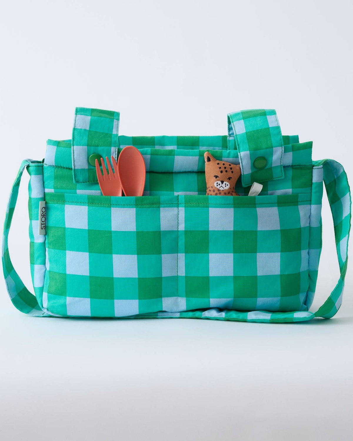 A green and white checkered picnic bag with handles and side straps, shown on a light gray background. The front pocket holds a coral-colored fork, a coral spoon, and a small plush toy.