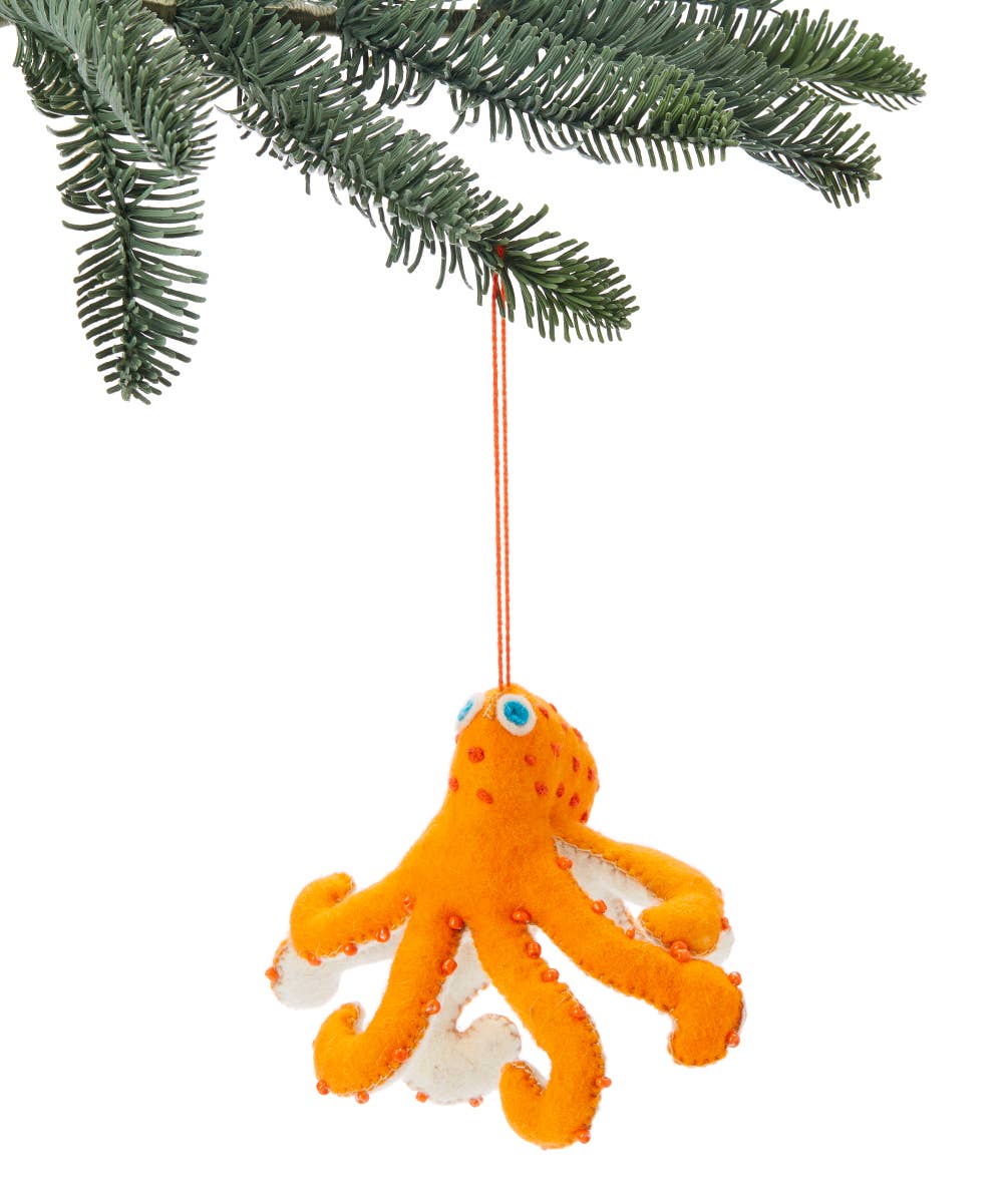 An orange felt octopus Christmas ornament with white tentacles and blue eyes, hanging from an orange string on a pine tree branch against a white background.