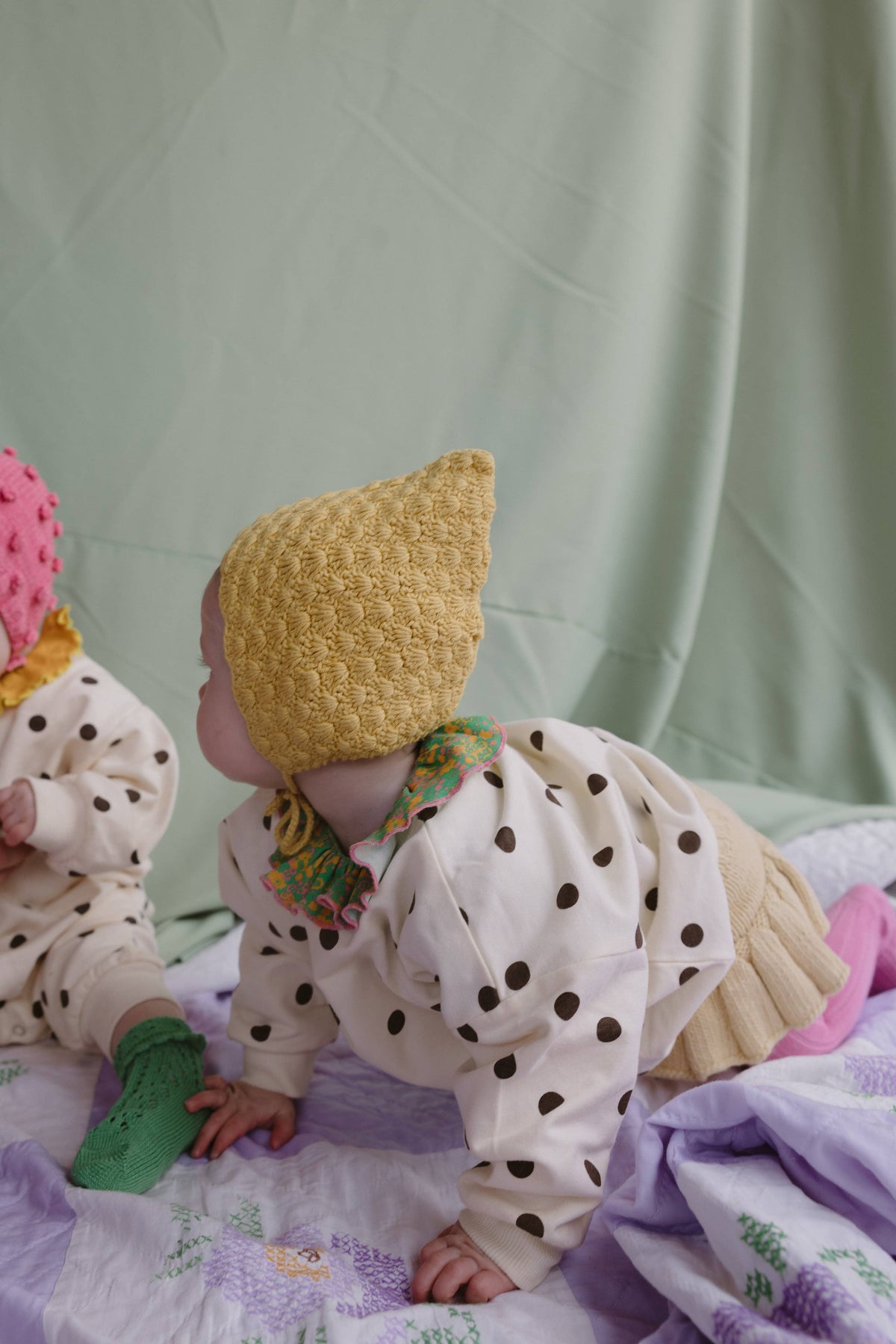 A baby crawling on a patterned quilt, wearing a mustard yellow knit pointed hat, a cream long-sleeve top with brown polka dots, and a beige skirt. Another baby in a pink hat is partially visible on the left against a sage green fabric backdrop.