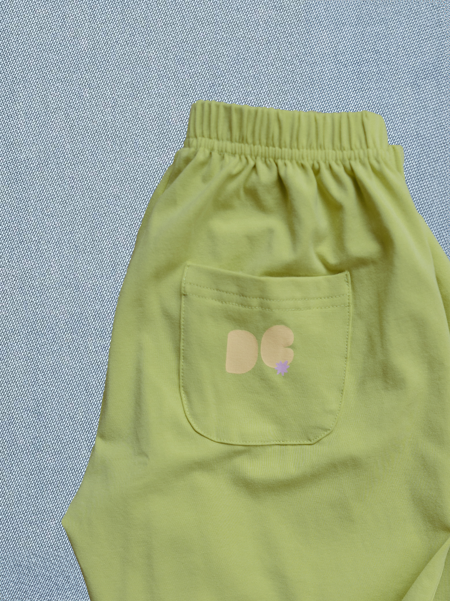 Light green pants with a pocket featuring a logo on a textured blue background