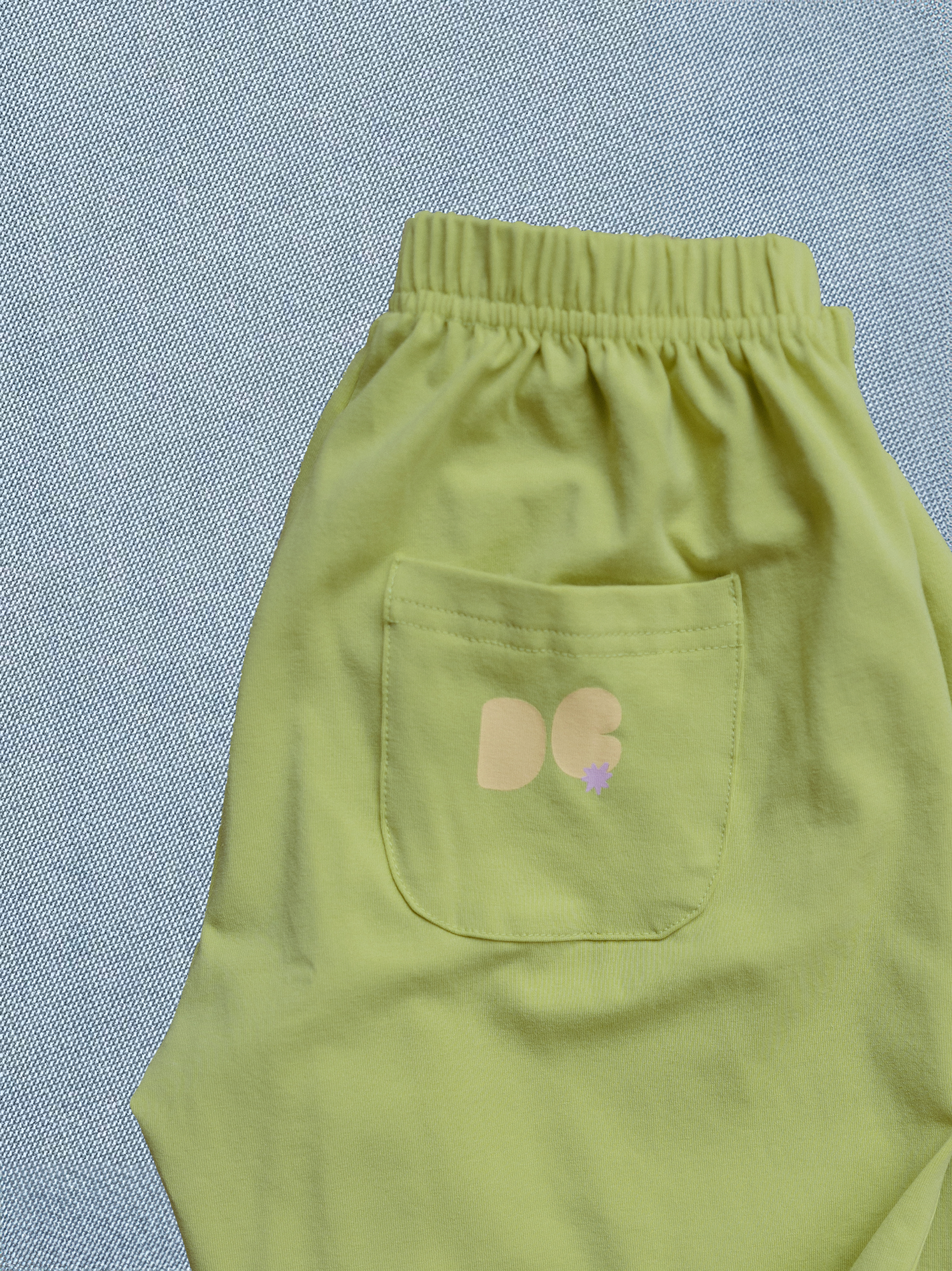 Light green pants with a pocket featuring a logo on a textured blue background