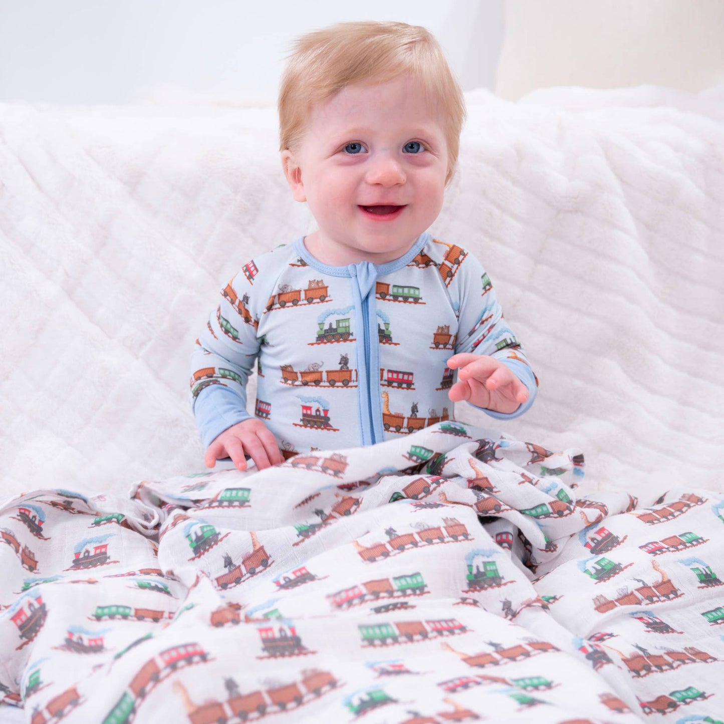 A smiling baby sitting on a white quilted bed, wearing a light blue zip-front sleeper with a brown and green train pattern, while covered with a matching train-print blanket.