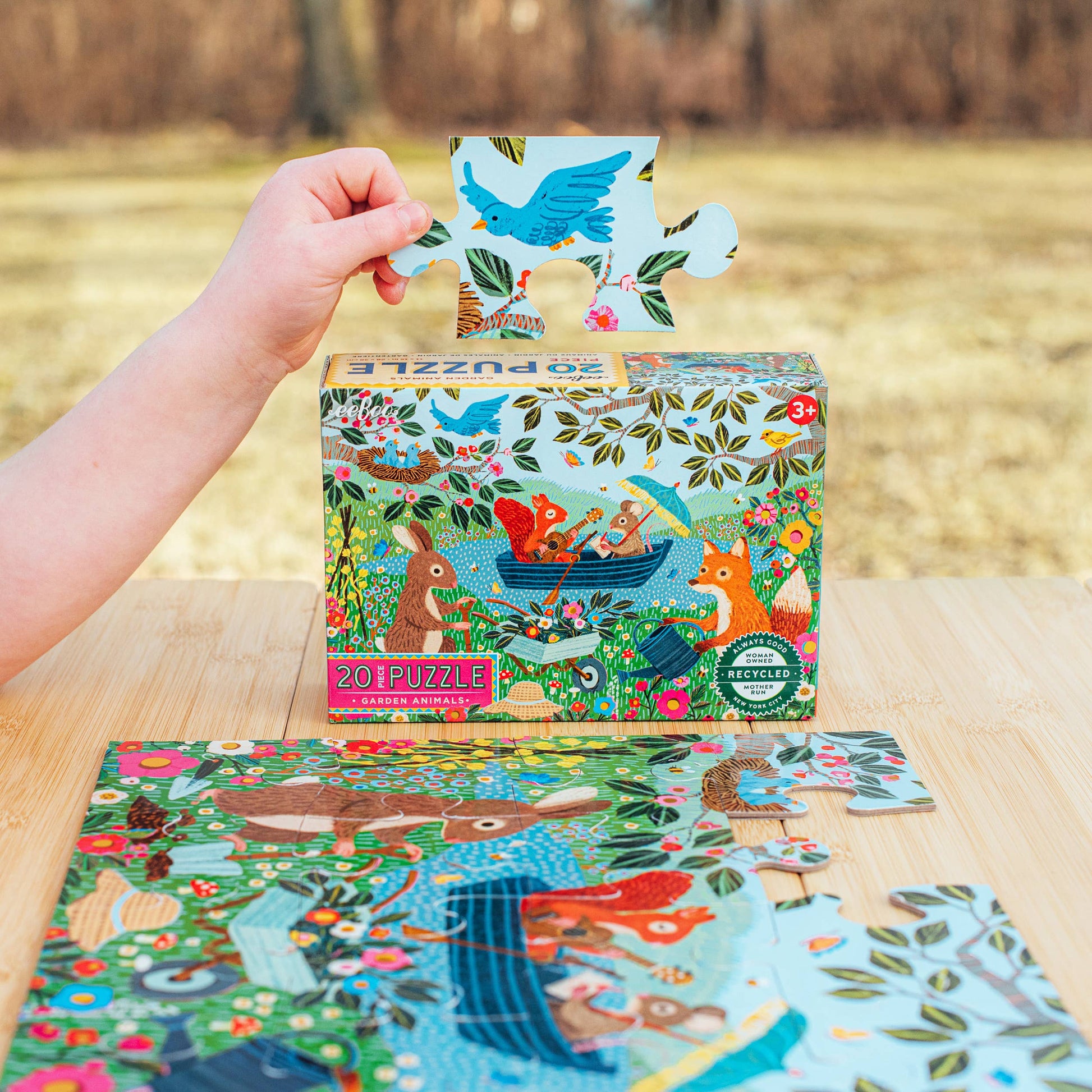 Child's hand holding a puzzle piece with a colorful puzzle box and completed puzzle on a wooden table.