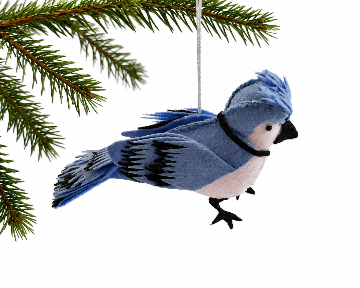 A blue and white felt bird ornament with black accents hangs from a white string beneath evergreen tree branches against a white background.
