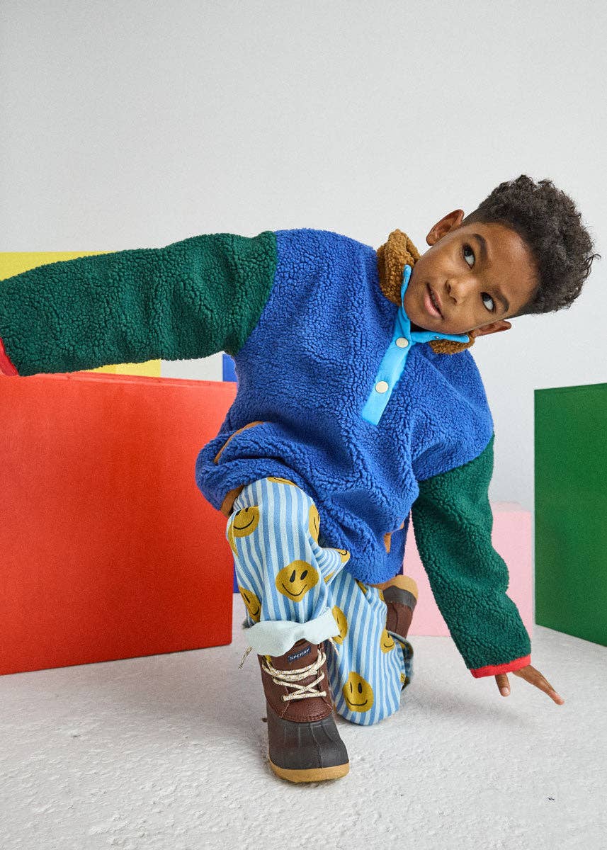 A young child wearing a colorful fleece pullover with green sleeves, a blue body, and a brown collar, paired with blue-and-white striped smiley-face pants and brown boots, posed in front of a white wall with colorful geometric blocks.