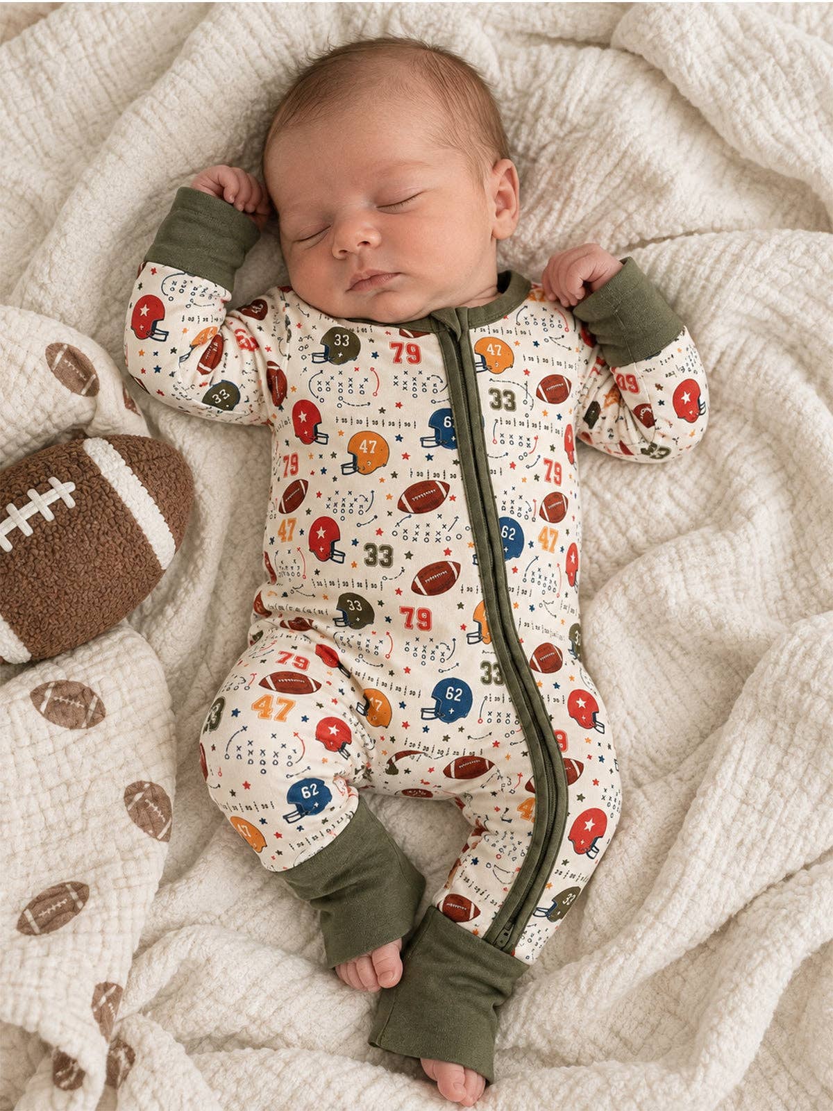 A sleeping newborn baby wearing a long-sleeve footed sleeper with an olive green zipper and cuffs, lying on a textured cream blanket next to a brown football.